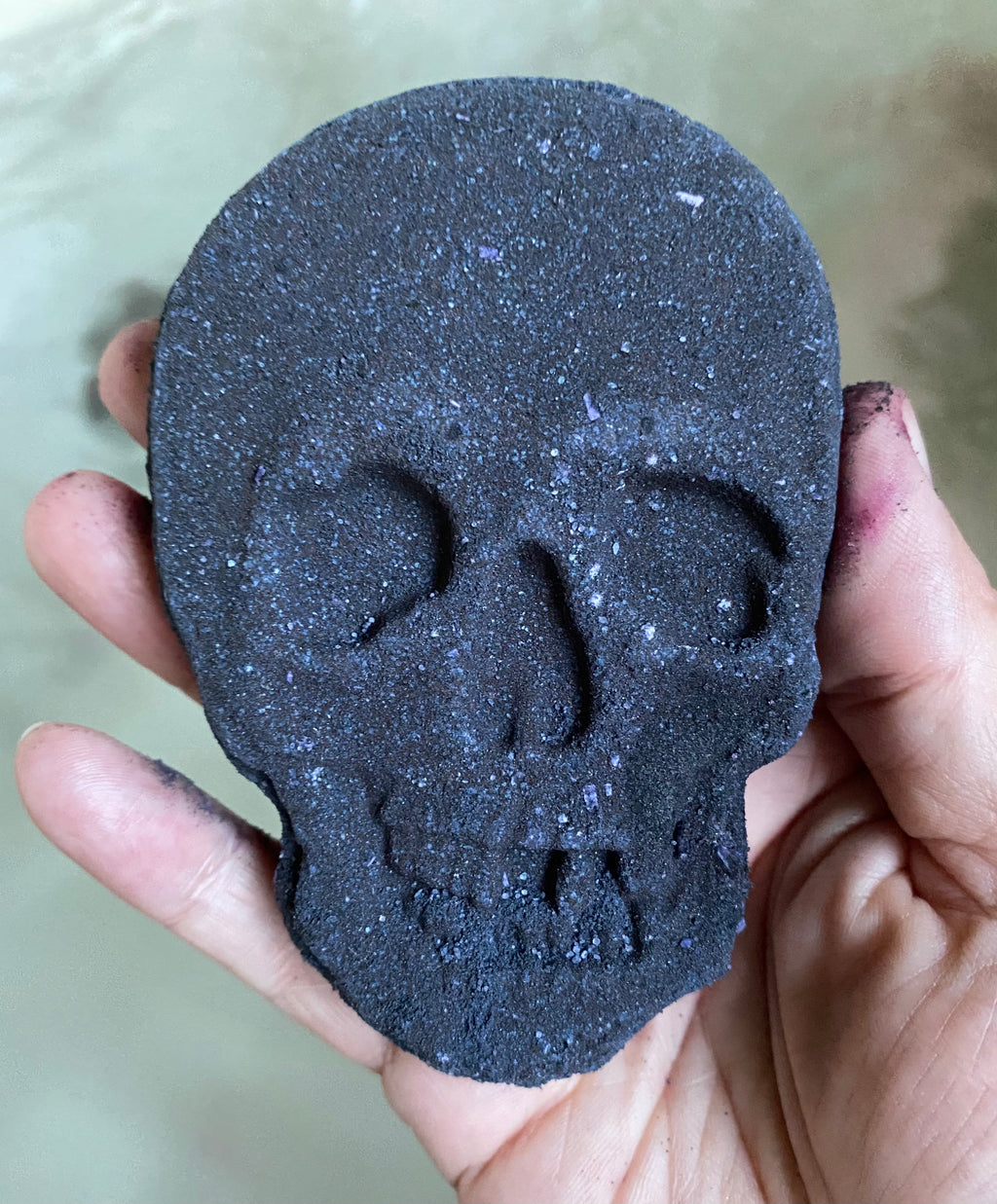 Skull shaped bath bomb in dark purple shown on the hand of the maker, above bath water.